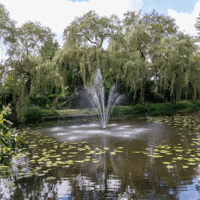 Decorative pond fountain spraying water among lily pads and weeping willow trees in a lush garden setting.