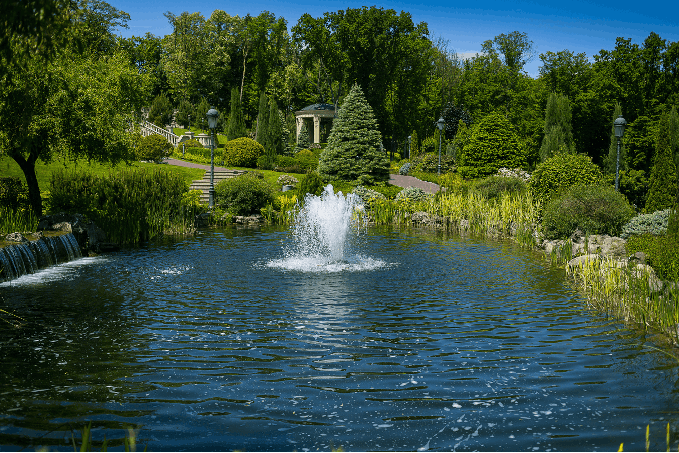 Decorative backyard pond with water fountain, small waterfall, manicured greenery, and gazebo in a landscaped garden setting