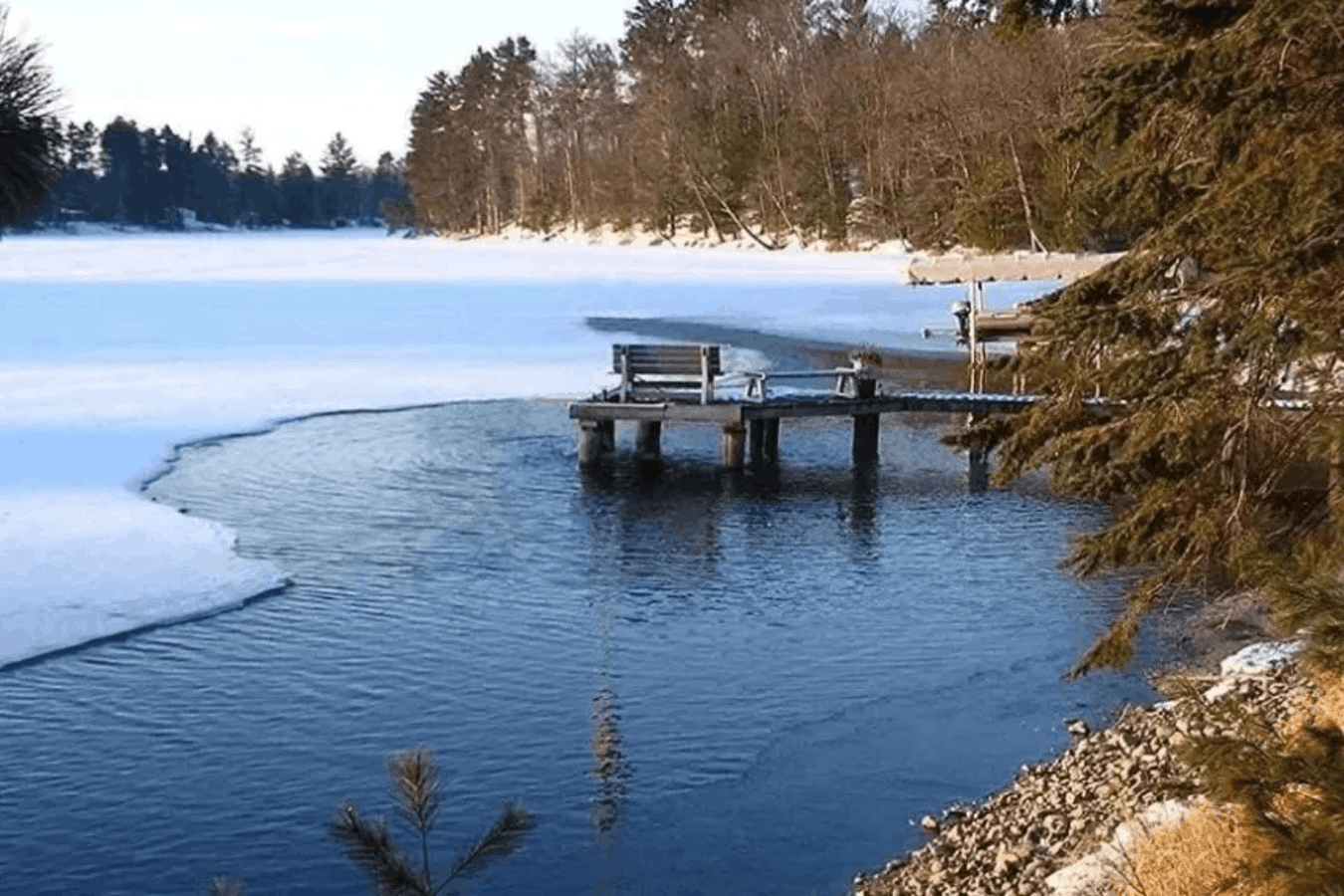 Partially frozen pond with open water near a wooden dock surrounded by snow and evergreen trees using a de-icer