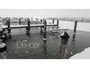 Snowy marina scene showing wooden docks and pilings partially covered in snow with an open water area around a de-icing device; snowflakes falling and calm water visible under overcast sky.