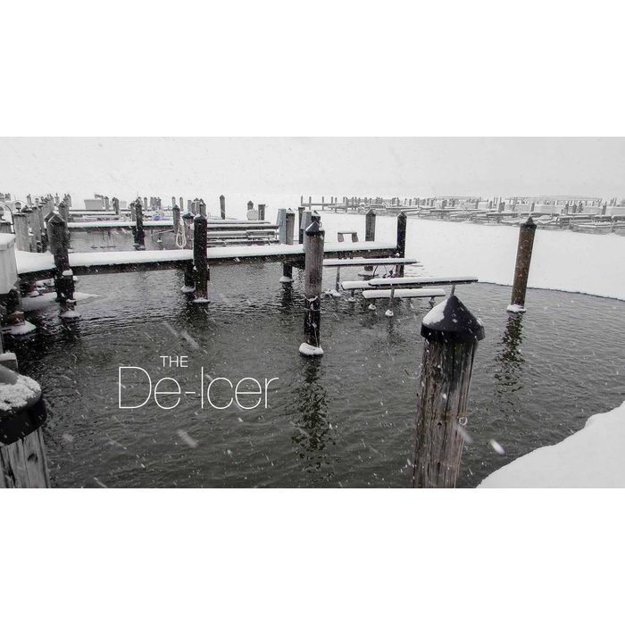 Snowy marina scene showing wooden docks and pilings partially covered in snow with an open water area around a de-icing device; snowflakes falling and calm water visible under overcast sky.